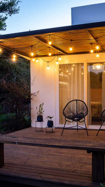 Alt text sugerido: Varanda moderna com piso de madeira, coberta por pergolado metálico com iluminação de lâmpadas pendentes. Duas cadeiras pretas e vasos de plantas decoram o espaço diante de uma porta de vidro com cortinas claras ao fundo, criando um ambiente aconchegante ao entardecer.