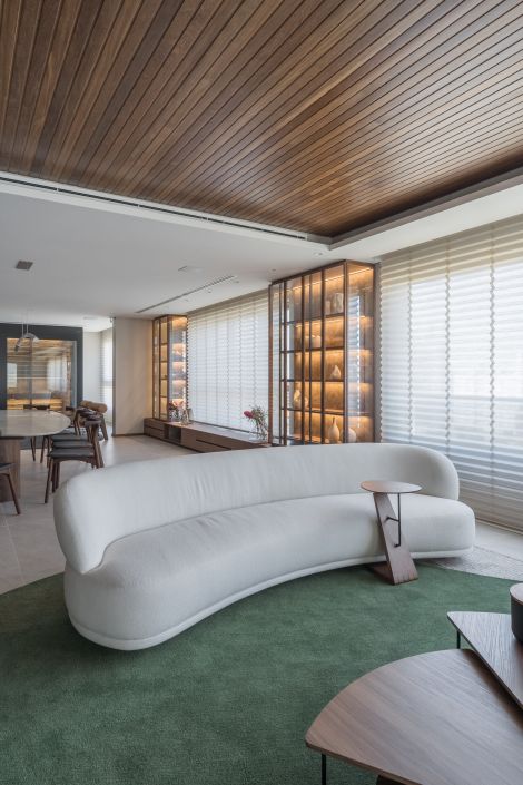 Sala de estar contemporânea com sofá curvo branco, tapete verde, cortinas com efeito ondulado e teto de madeira, em ambiente integrado com sala de jantar. Projeto do Studio Colnaghi.