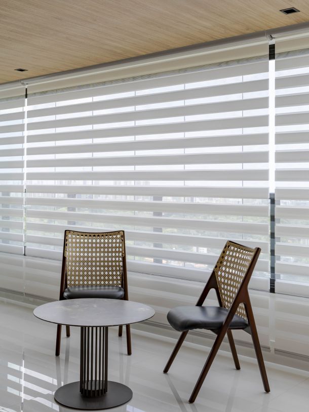 Estúdio da arquiteta com cortina Mystique Mutabile, duas cadeiras de madeira e palhinha e mesa de apoio sobre piso claro, em ambiente iluminado e contemporâneo.