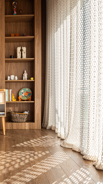 Ambiente com piso de madeira e cortinas de trama rendada que filtram a luz natural, projetando sombras no chão. Ao lado, uma estante de madeira com nichos decorados por livros, esculturas e objetos coloridos compõe um espaço aconchegante e cheio de personalidade.