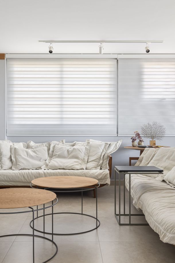 Sala de estar clara e acolhedora com cortina Mystique da Uniflex, especificada pela loja Uniflex Vitória Decor e pelo escritório Mutabile. O ambiente possui sofás em linho natural, mesas de centro leves e tons neutros, iluminado por luz suave filtrada pela cortina.