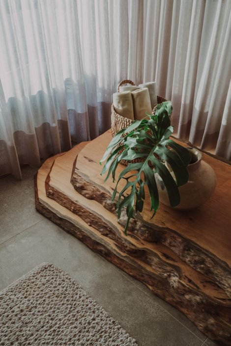 Detalhe de ambiente com cortina translúcida, mesa de madeira rústica com bordas naturais, vaso de cerâmica com folha de costela-de-adão e cesto de palha com mantas. Projeto do Panazzolo Estúdio.