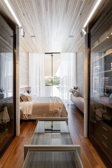 Fachada com amplas portas de vidro e cortinas brancas translúcidas, revelando um quarto com teto e piso de madeira. O ambiente reflete elegância e integração com o exterior.