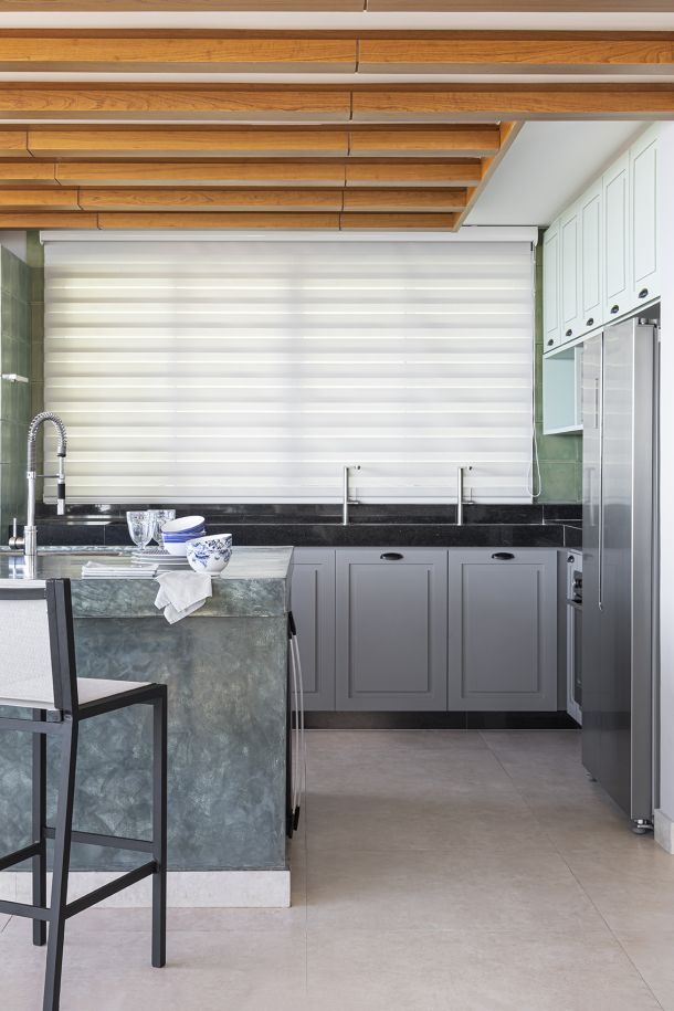 Cozinha moderna com cortina Mutabile da Vitória Decor, bancada verde, armários acinzentados e teto com vigas de madeira aparente.
