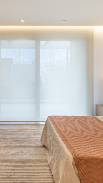 Sala de jantar com mesa oval de madeira escura, cadeiras em couro caramelo e tapete colorido. Ao fundo, cortinas de tecido leve em tom neutro filtram a luz natural, criando um ambiente elegante e acolhedor com detalhes em madeira nas paredes.