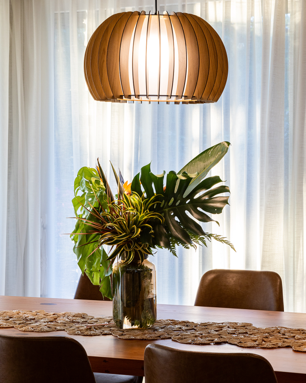 Close-up de mesa de jantar em madeira com arranjo de folhas tropicais em vaso de vidro. Ao fundo, cortinas brancas filtram a luz natural, e uma luminária pendente em ripas de madeira ilumina o ambiente, criando uma atmosfera acolhedora e elegante.