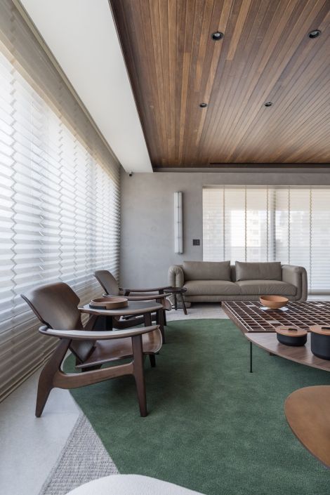 Sala de estar com teto de madeira, cortinas em tecido leve com efeito ondulado, poltronas e mesa de centro de madeira, sofá em tom neutro e tapete verde. Projeto do Studio Colnaghi.