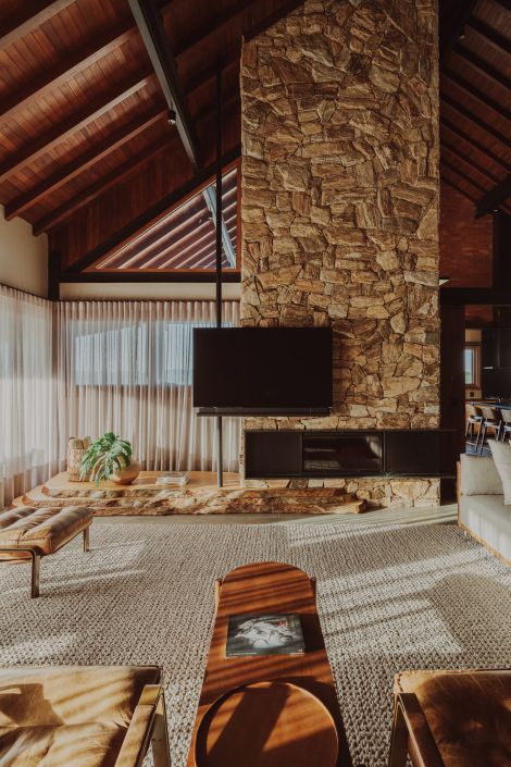 Sala de estar com parede de pedra natural, teto de madeira com vigas aparentes, cortinas translúcidas e mobiliário em tons terrosos, criando um ambiente aconchegante e sofisticado.