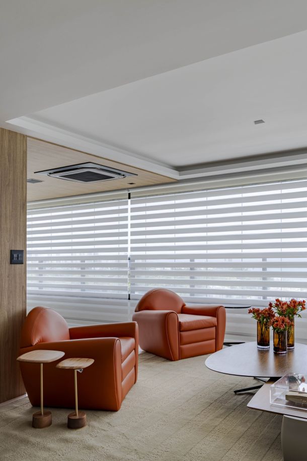 Sala contemporânea com cortina Mystique da Uniflex, assinada pela arquiteta Daniela Lopes. O ambiente combina tons neutros e poltronas em couro alaranjado, com iluminação natural filtrada pela cortina, criando uma atmosfera moderna e acolhedora.