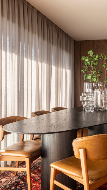 Sala de jantar com mesa oval de madeira escura, cadeiras em couro caramelo e tapete colorido. Ao fundo, cortinas de tecido leve em tom neutro filtram a luz natural, criando um ambiente elegante e acolhedor com detalhes em madeira nas paredes.