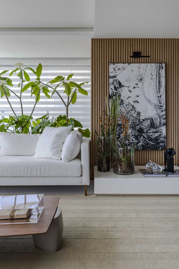 Sala de estar elegante e natural com cortina Mystique da Uniflex, assinada pela arquiteta Daniela Lopes. O ambiente combina sofá branco, parede ripada de madeira e plantas tropicais, com iluminação suave filtrada pela cortina.