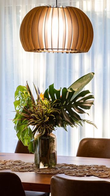 Close-up de mesa de jantar em madeira com arranjo de folhas tropicais em vaso de vidro. Ao fundo, cortinas brancas filtram a luz natural, e uma luminária pendente em ripas de madeira ilumina o ambiente, criando uma atmosfera acolhedora e elegante.