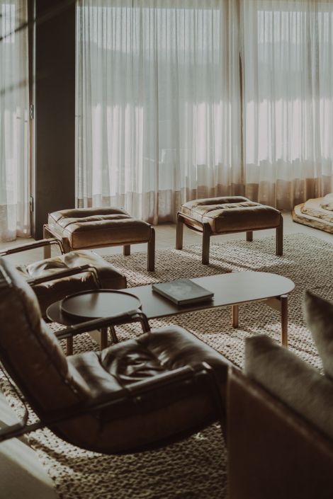 Sala de estar com poltronas e pufes de couro, cortinas translúcidas em tecido leve e iluminação natural suave, criando atmosfera acolhedora e sofisticada. Projeto do Panazzolo Estúdio.