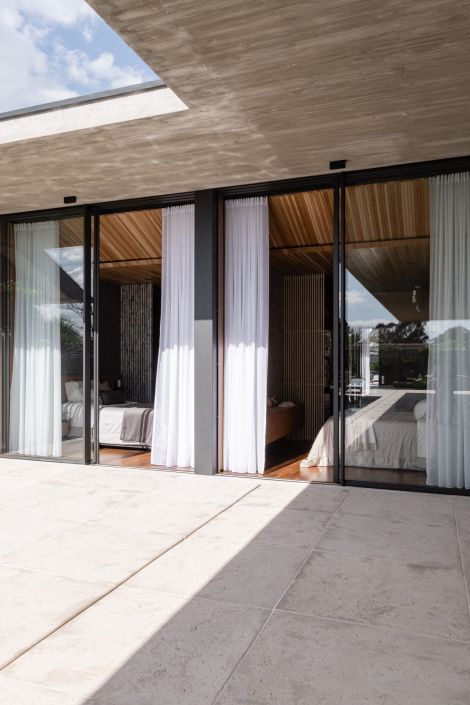 Fachada com amplas portas de vidro e cortinas brancas translúcidas, revelando um quarto com teto e piso de madeira. O ambiente reflete elegância e integração com o exterior.