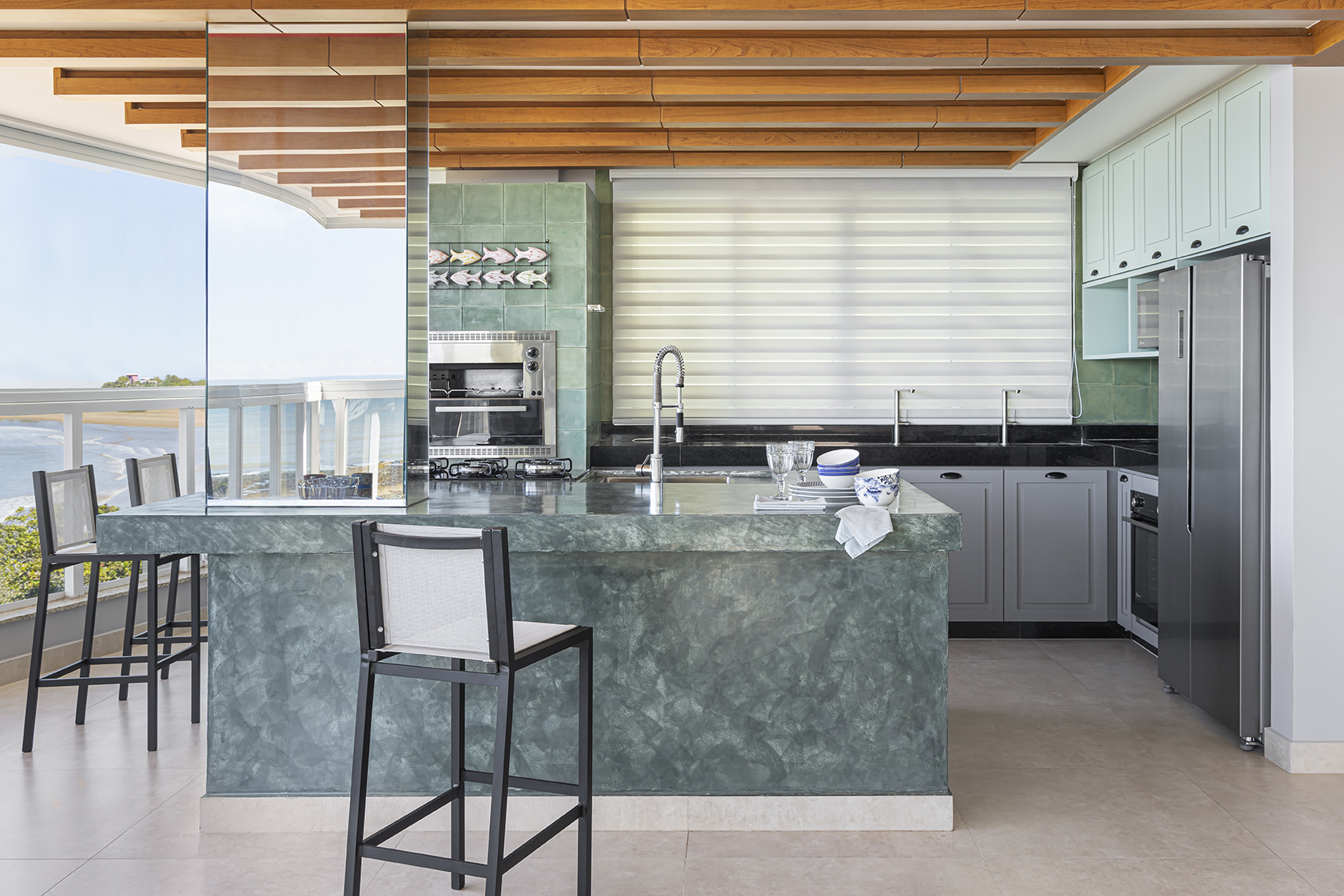 Cozinha sofisticada com cortina Mutabile da Vitória Decor, bancada em tom verde, detalhes em madeira e vista aberta para o mar.