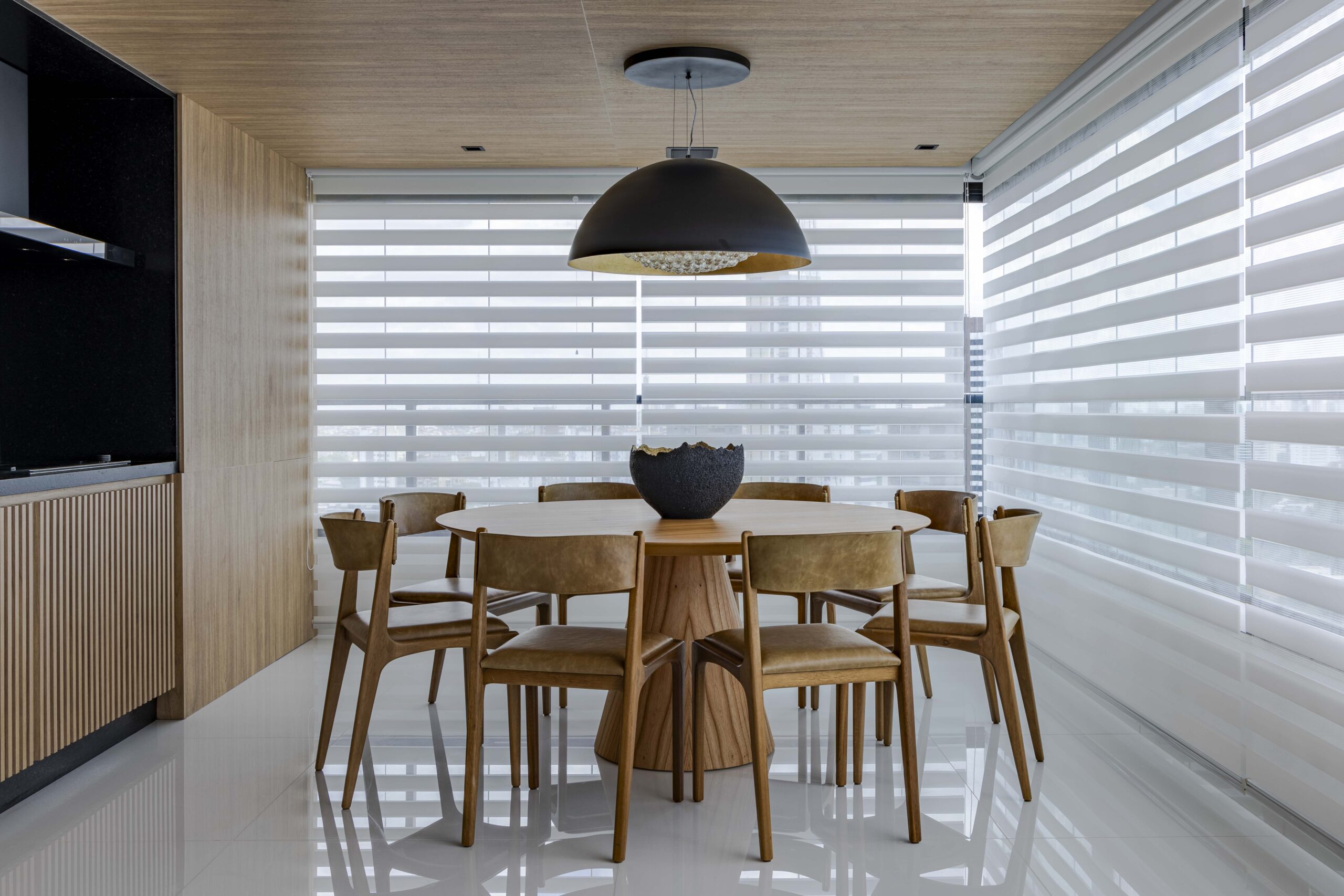 Sala de jantar moderna com cortinas Paradise da Trilho Suisso, filtrando a luz natural através de faixas horizontais translúcidas, mesa redonda de madeira e cadeiras estofadas em couro bege.