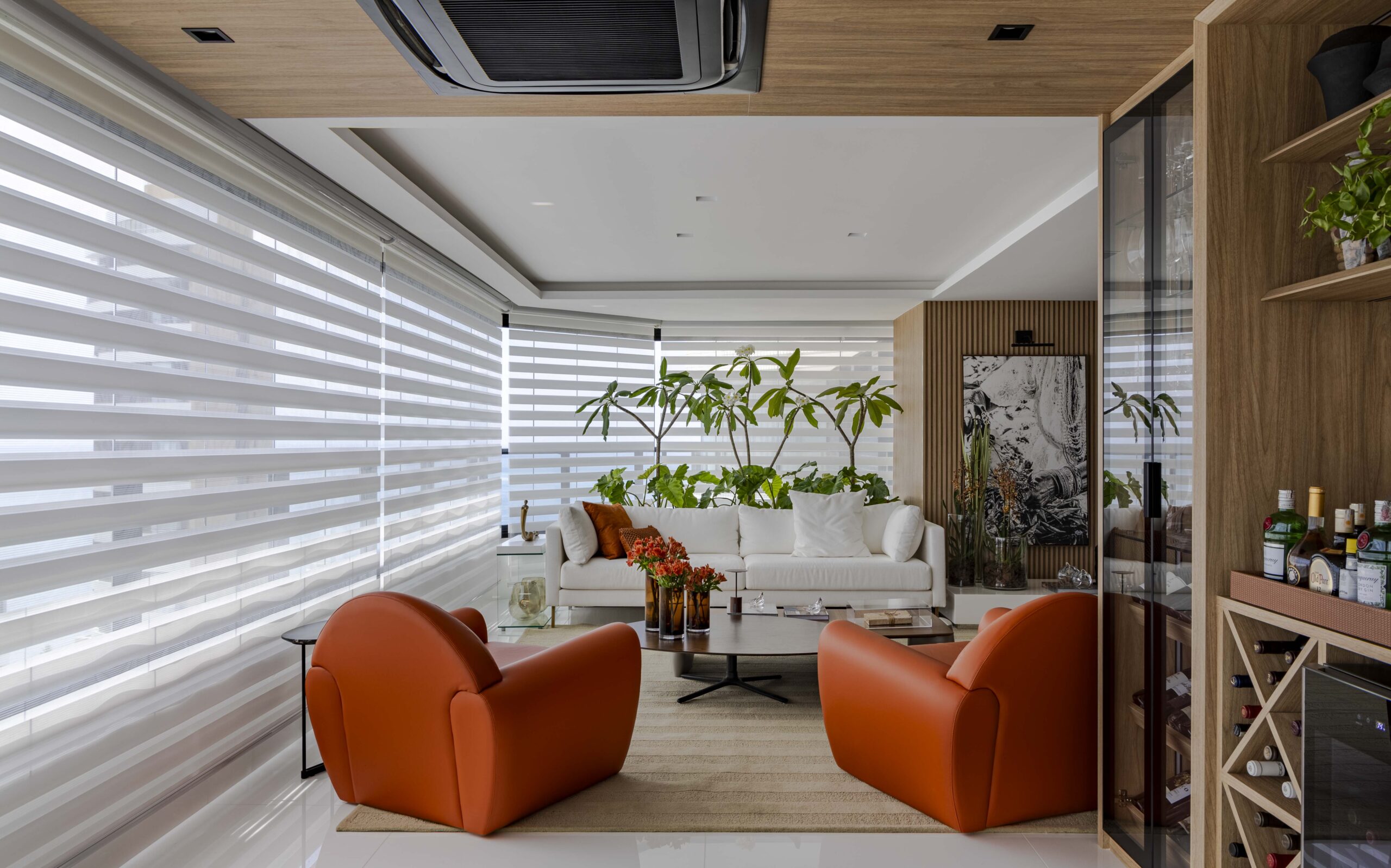 Sala de estar contemporânea com cortinas Paradise da Trilho Suisso, sofá branco, poltronas laranja e plantas naturais ao fundo, em ambiente iluminado e elegante.